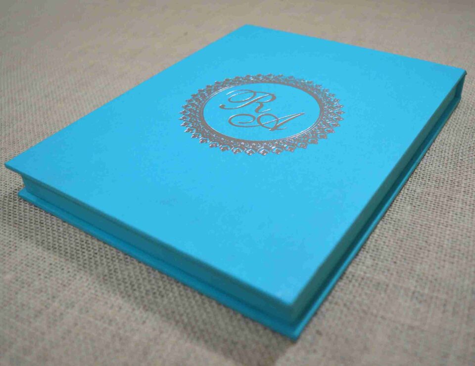 An angled side view of a closed, rectangular turquoise silk invitation box resting on a textured, neutral-toned fabric surface. The flat lid is centered with a shimmering silver foil monogram featuring the initials in an elegant script. The initials are enclosed within a circular, decorative silver border with a delicate, lace-like pattern. The clean lines of the box and the vibrant turquoise color give the invitation a modern yet sophisticated appearance.
