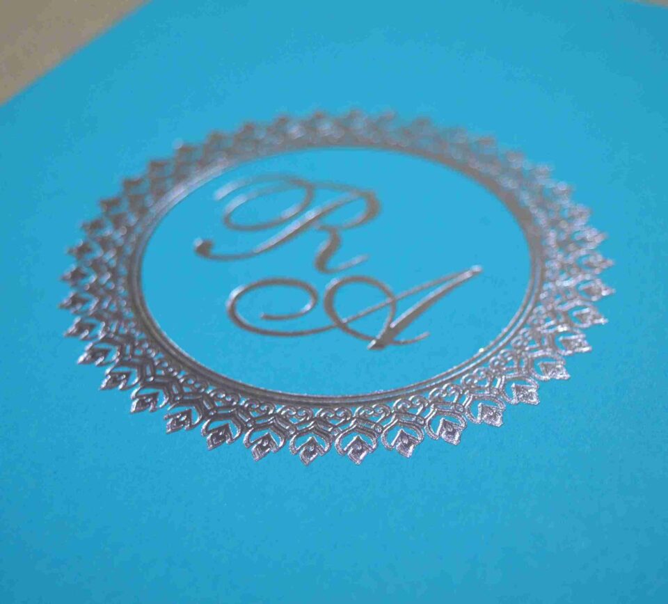 An angled side view of a closed, rectangular turquoise silk invitation box resting on a textured, neutral-toned fabric surface. The flat lid is centered with a shimmering silver foil monogram featuring the initials in an elegant script. The initials are enclosed within a circular, decorative silver border with a delicate, lace-like pattern. The clean lines of the box and the vibrant turquoise color give the invitation a modern yet sophisticated appearance.