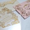 An ornate wedding invitation suite is displayed on a white surface, featuring ivory, pink, and transparent acrylic elements decorated with elaborate gold foil filigree.