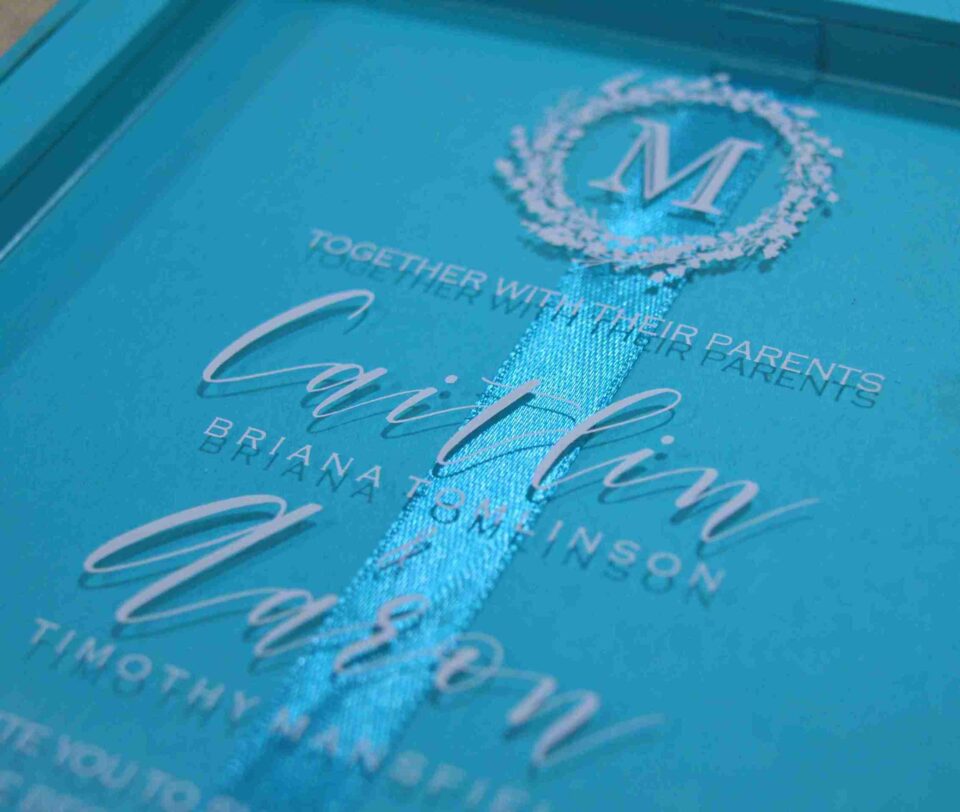 A high-angle view of an open turquoise silk invitation box resting on a textured, neutral-toned fabric surface.The presentation includes:The left panel: Features a large, ornate silver Logo encrusted with sparkling crystals.The center A clear acrylic invitation card sits within the folio, featuring the name in a delicate white script, along with formal wedding details.The right panel: Contains a pocket holding coordinating stationery, including a white RSVP, card with a silver-foiled decorative border. The overall design is vibrant and modern, accented by the shimmering silver elements.