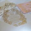 An ornate wedding invitation suite is displayed on a white surface, featuring ivory, pink, and transparent acrylic elements decorated with elaborate gold foil filigree.