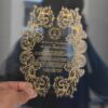 A hand holds up a transparent acrylic wedding invitation with an intricate, laser-cut scalloped edge. The invitation is decorated with a heavy gold foil filigree border of swirling acanthus leaves that frame the central text.