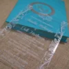 A high-angle view of an open turquoise silk invitation box resting on a textured, neutral-toned fabric surface.The presentation includes:The left panel: Features a large, ornate silver Logo encrusted with sparkling crystals.The center A clear acrylic invitation card sits within the folio, featuring the name in a delicate white script, along with formal wedding details.The right panel: Contains a pocket holding coordinating stationery, including a white RSVP, card with a silver-foiled decorative border. The overall design is vibrant and modern, accented by the shimmering silver elements.
