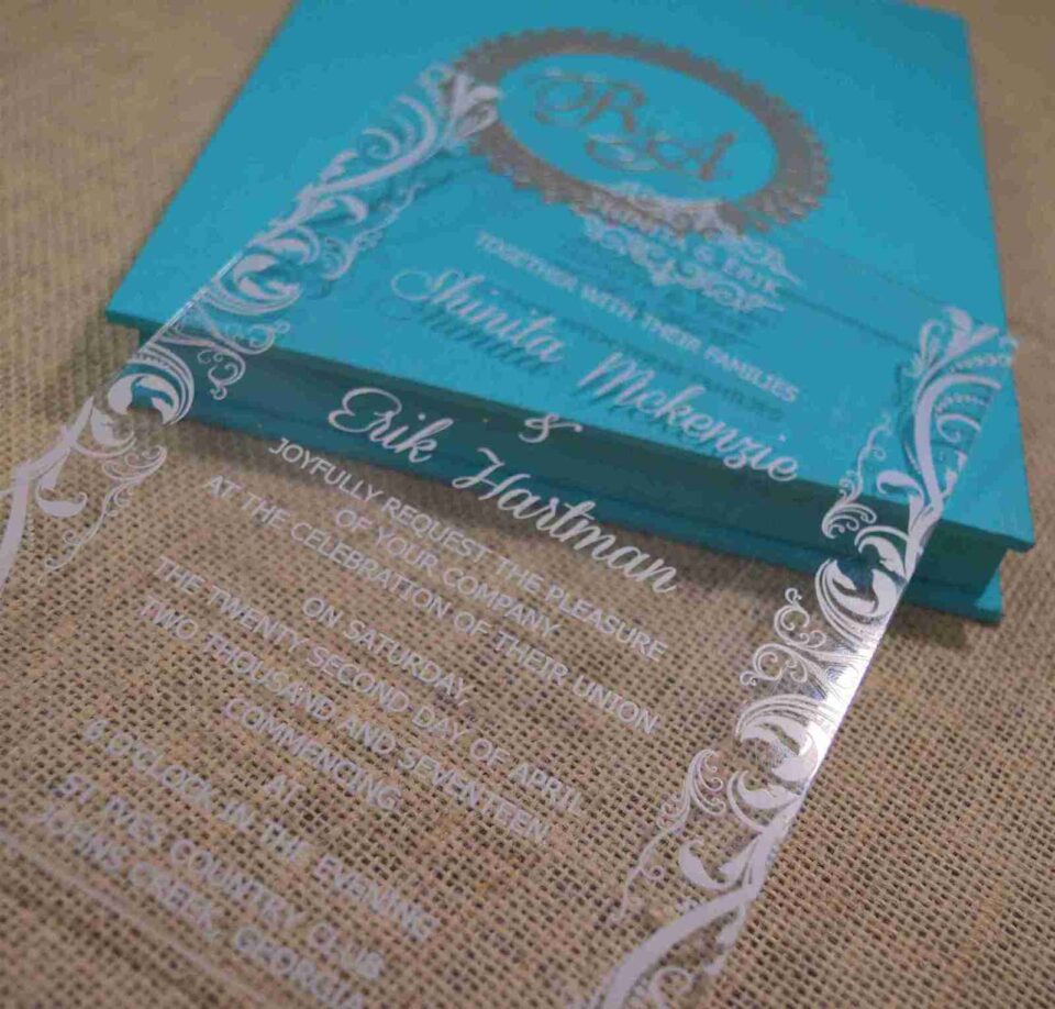 A high-angle view of an open turquoise silk invitation box resting on a textured, neutral-toned fabric surface.The presentation includes:The left panel: Features a large, ornate silver Logo encrusted with sparkling crystals.The center A clear acrylic invitation card sits within the folio, featuring the name in a delicate white script, along with formal wedding details.The right panel: Contains a pocket holding coordinating stationery, including a white RSVP, card with a silver-foiled decorative border. The overall design is vibrant and modern, accented by the shimmering silver elements.