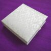 A closed, square white invitation box covered in an elegant lace floral pattern. The box is shown at an angle, resting on a textured purple fabric background.