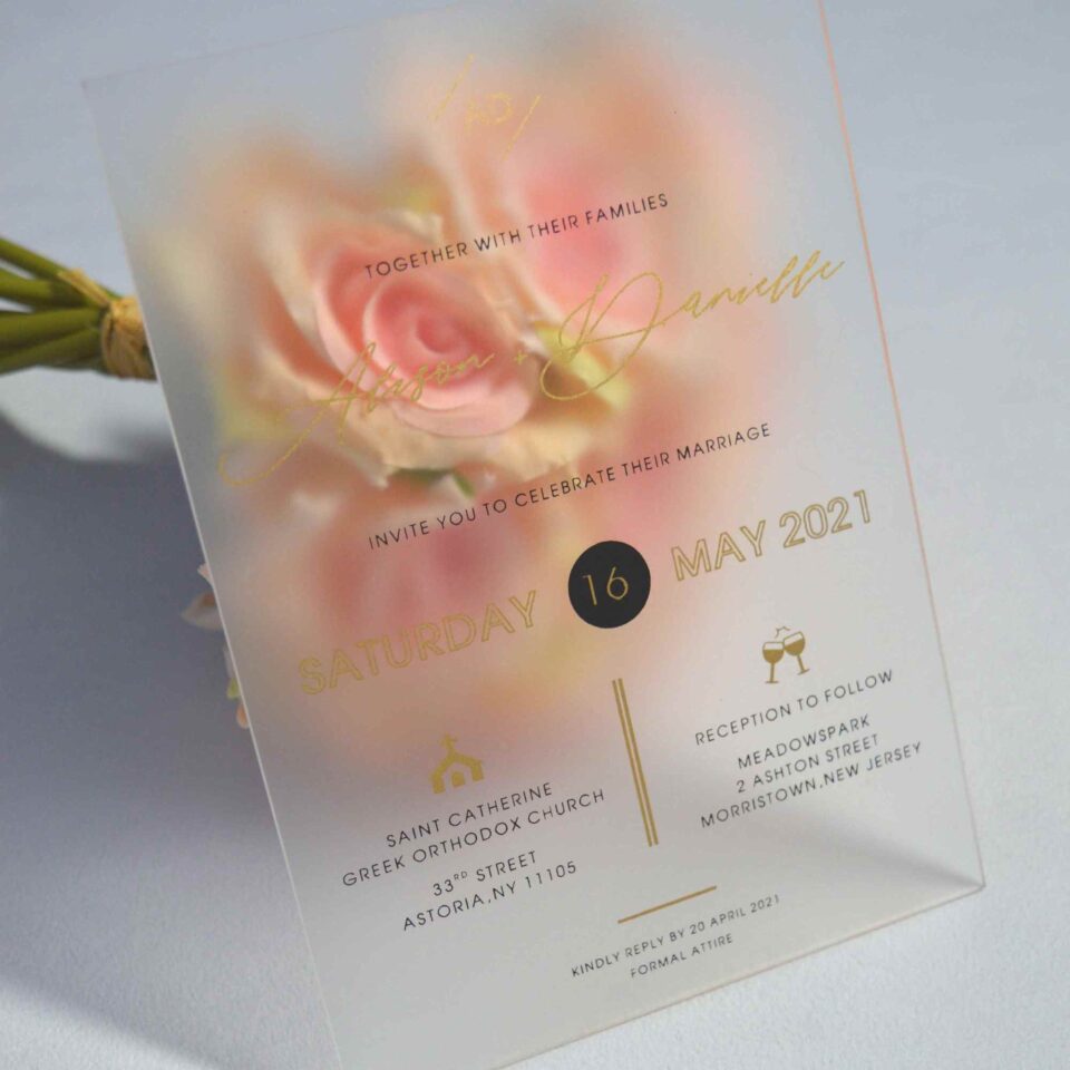 An elegant Frosted acrylic wedding invitation is shown leaning against a lush bouquet of soft peach and pink roses on a white surface. The invitation features a minimalist design with gold foil text and a delicate gold geometric line pattern at the bottom. The text on the clear card includes Details