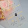 An elegant Frosted acrylic wedding invitation is shown leaning against a lush bouquet of soft peach and pink roses on a white surface. The invitation features a minimalist design with gold foil text and a delicate gold geometric line pattern at the bottom. The text on the clear card includes Details