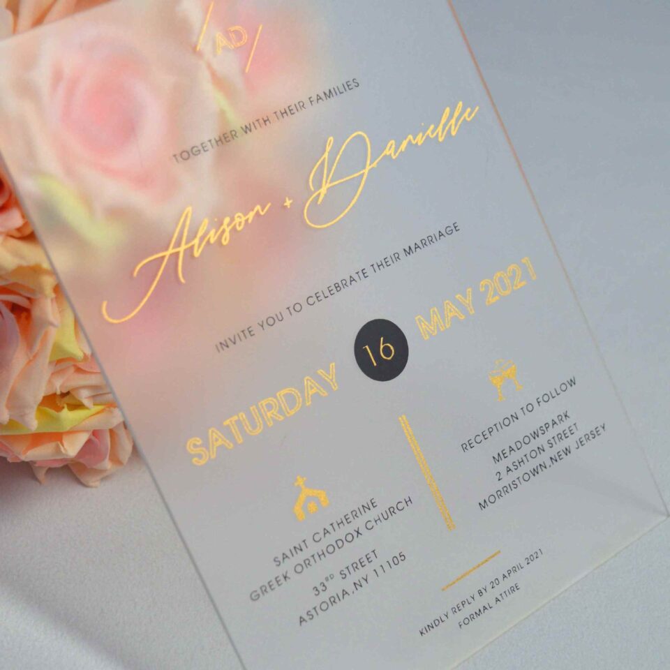 An elegant Frosted acrylic wedding invitation is shown leaning against a lush bouquet of soft peach and pink roses on a white surface. The invitation features a minimalist design with gold foil text and a delicate gold geometric line pattern at the bottom. The text on the clear card includes Details