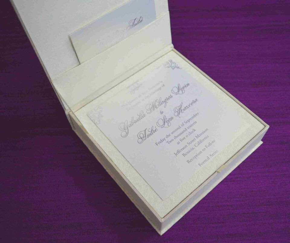 Here is the ALT text for the final image in your collection: **DSC_0293_165_11zon-960x804.jpg** An open, square ivory-colored wedding invitation box resting on a textured purple fabric surface. The interior lid features a horizontal pocket containing a small card for "Gabriella and Andre." The base of the box holds the main wedding invitation for "Gabriella Milagros Levin and Andre Leon Keavera," printed in elegant script on a white card with delicate corner flourishes.