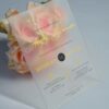 An elegant Frosted acrylic wedding invitation is shown leaning against a lush bouquet of soft peach and pink roses on a white surface. The invitation features a minimalist design with gold foil text and a delicate gold geometric line pattern at the bottom. The text on the clear card includes Details