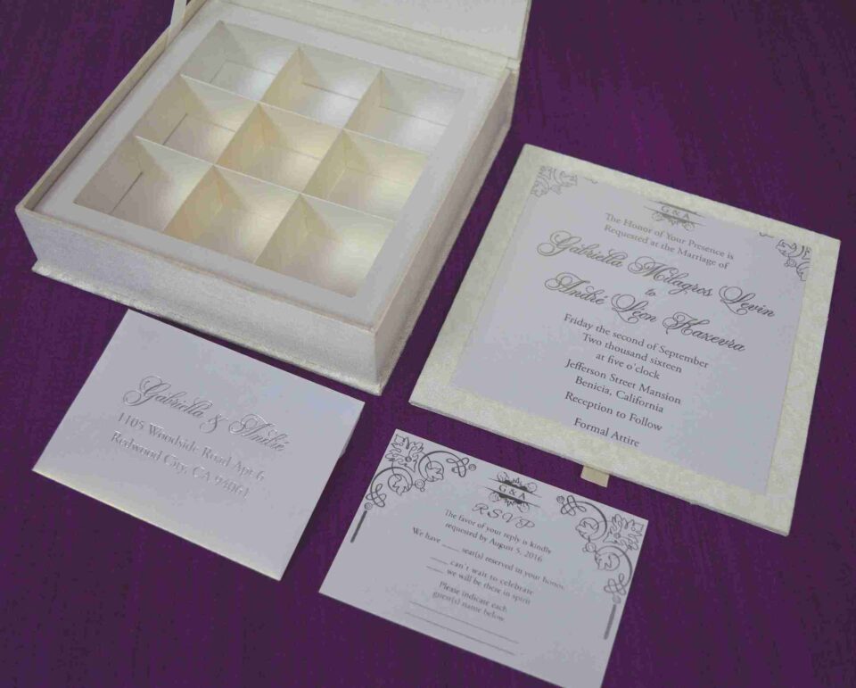 Here is the ALT text for the final image in your collection: **DSC_0294_161_11zon-960x706.jpg** An open, ivory-colored invitation box set against a textured purple background. The interior of the lid features a horizontal pocket holding a matching name card. The base of the box contains a white wedding invitation for "Gabriella Milagros Levin and Andre Leon Keavera," which is tilted to reveal a recessed grid with six empty compartments underneath, designed for holding favors or chocolates.