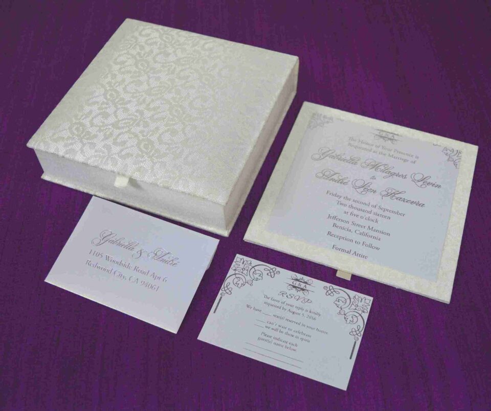 Here is the ALT text for the final image in your collection: **DSC_0293_165_11zon-960x804.jpg** An open, square ivory-colored wedding invitation box resting on a textured purple fabric surface. The interior lid features a horizontal pocket containing a small card for "Gabriella and Andre." The base of the box holds the main wedding invitation for "Gabriella Milagros Levin and Andre Leon Keavera," printed in elegant script on a white card with delicate corner flourishes.