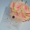 An elegant Frosted acrylic wedding invitation is shown leaning against a lush bouquet of soft peach and pink roses on a white surface. The invitation features a minimalist design with gold foil text and a delicate gold geometric line pattern at the bottom. The text on the clear card includes Details