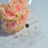 A transparent acrylic wedding invitation is displayed at an angle, leaning against a bouquet of soft peach-colored roses. The invitation features elegant gold text and icons that appear to float over the flowers visible through the clear material.
