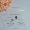 A transparent acrylic wedding invitation is displayed at an angle, leaning against a bouquet of soft peach-colored roses. The invitation features elegant gold text and icons that appear to float over the flowers visible through the clear material.
