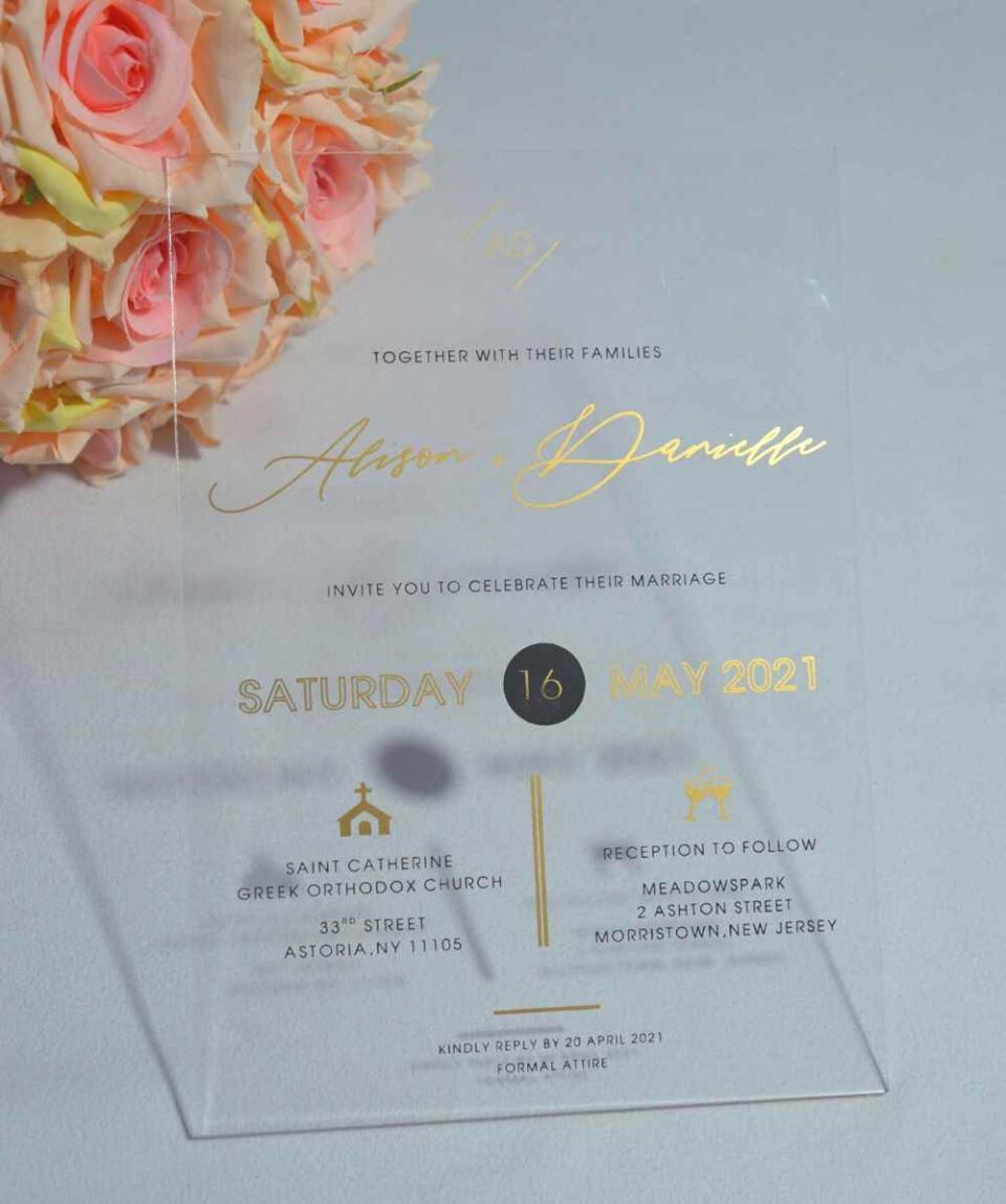 A transparent acrylic wedding invitation is displayed at an angle, leaning against a bouquet of soft peach-colored roses. The invitation features elegant gold text and icons that appear to float over the flowers visible through the clear material.