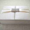 A top-down view of a square, champagne-colored silk invitation box resting on a textured white surface. A wide, matching champagne satin ribbon is wrapped horizontally across the lid, featuring a large, circular gold brooch at the center. The brooch is elaborately designed with concentric rings of shimmering clear crystals. A few loose pink flower petals are scattered around the box, and a stem of vibrant pink gladiolus flowers is partially visible on the right.
