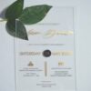 A transparent acrylic wedding invitation is displayed at an angle, leaning against a bouquet of soft green leaves resting over the top left corner.The invitation features elegant gold text.