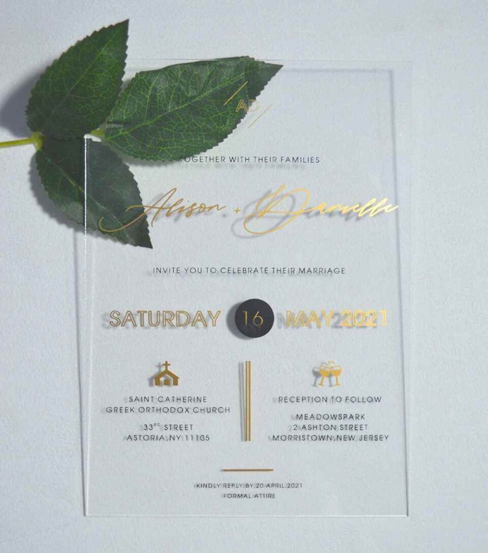A transparent acrylic wedding invitation is displayed at an angle, leaning against a bouquet of soft green leaves resting over the top left corner.The invitation features elegant gold text.