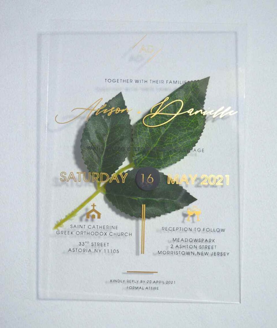 A transparent acrylic wedding invitation is displayed at an angle, leaning against a bouquet of soft green leaves resting over the top left corner.The invitation features elegant gold text.