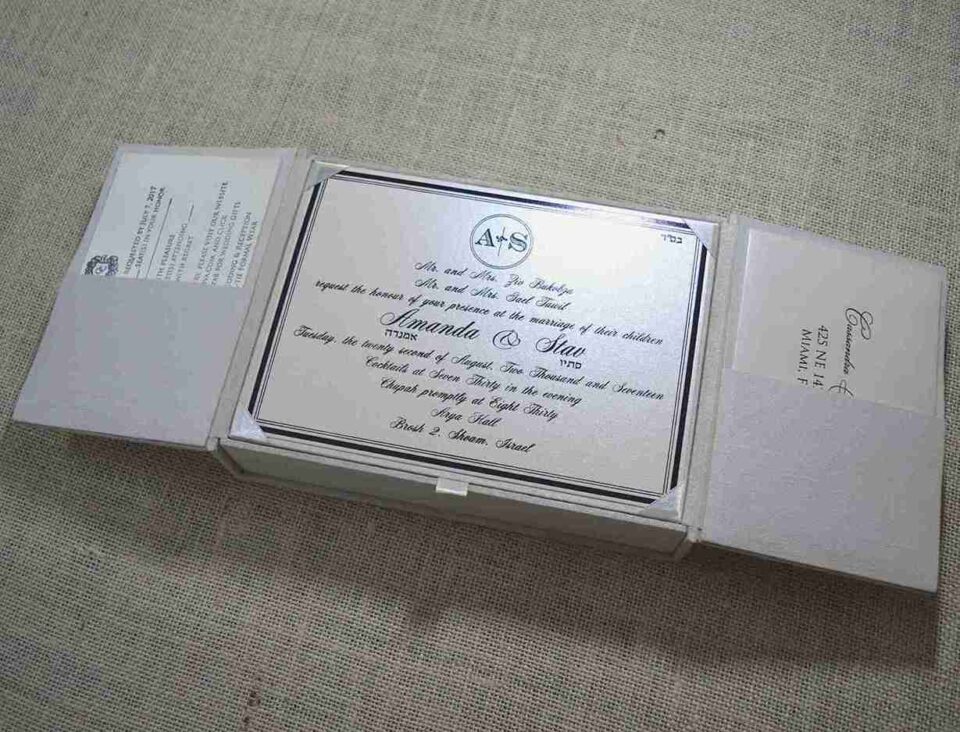 A top-down view of an open, champagne-colored silk invitation box. The interior lid features a matching horizontal silk pocket holding a white card printed with the name "Mathew and Michelle" in elegant gold script. The base of the box contains a formal wedding invitation card with gold-foil floral accents in the corners. A stem of vibrant pink gladiolus flowers lies to the right of the box on a textured white surface.
