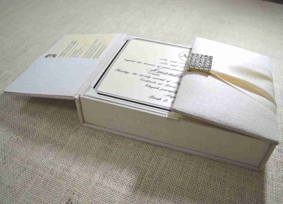 A top-down view of an open, champagne-colored silk invitation box. The interior lid features a matching horizontal silk pocket holding a white card printed with the name "Mathew and Michelle" in elegant gold script. The base of the box contains a formal wedding invitation card with gold-foil floral accents in the corners. A stem of vibrant pink gladiolus flowers lies to the right of the box on a textured white surface.