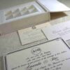 A flat-lay of a white wedding gift suite. It features a closed box with a crystal brooch, an open box base containing nine gold-wrapped chocolate truffles in a grid, a formal invitation for Mathew and Michelle, an RSVP card, and an envelope, all accented by pink flowers.