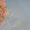 An transparent acrylic wedding invitation resting on a white surface. The card is leaning against a bouquet of soft peach and pink roses. The invitation features minimalist gold foil text with details.
