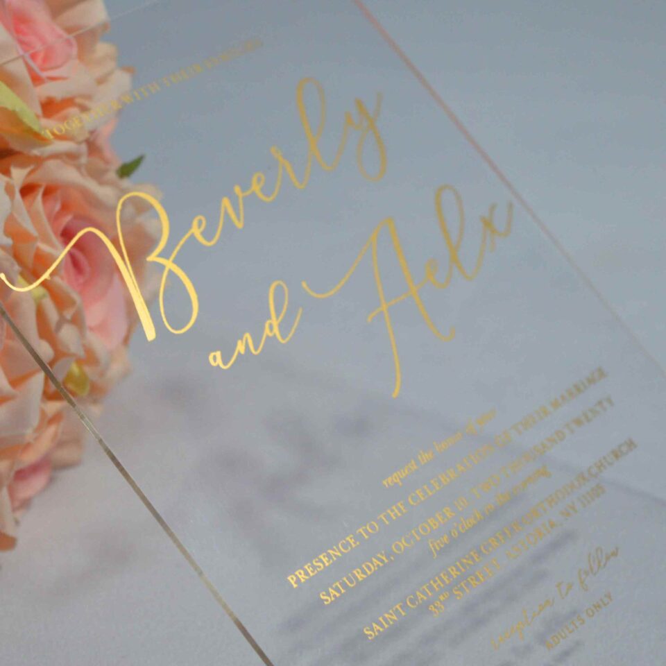 An transparent acrylic wedding invitation resting on a white surface. The card is leaning against a bouquet of soft peach and pink roses. The invitation features minimalist gold foil text with details.