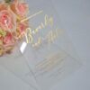 An transparent acrylic wedding invitation resting on a white surface. The card is leaning against a bouquet of soft peach and pink roses. The invitation features minimalist gold foil text with details.