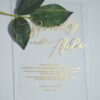 A transparent, rectangular acrylic wedding invitation is laid flat on a white surface, accompanied by a stem of green leaves at the top left. The invitation features elegant gold text that casts subtle shadows onto the background.