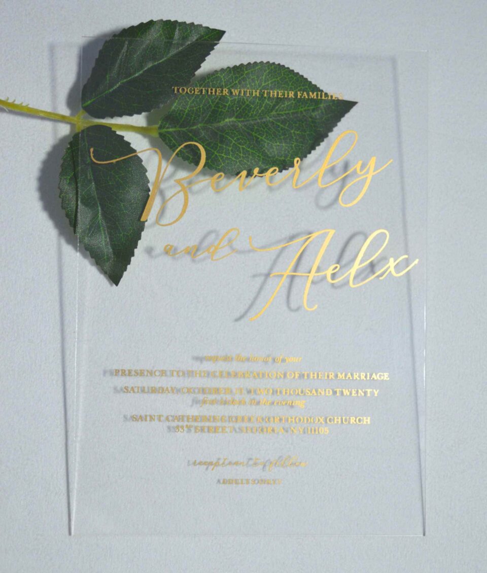 A transparent, rectangular acrylic wedding invitation is laid flat on a white surface, accompanied by a stem of green leaves at the top left. The invitation features elegant gold text that casts subtle shadows onto the background.