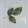 A transparent, rectangular acrylic wedding invitation is laid flat on a white surface, accompanied by a stem of green leaves at the center. The invitation features elegant gold text that casts subtle shadows onto the background.