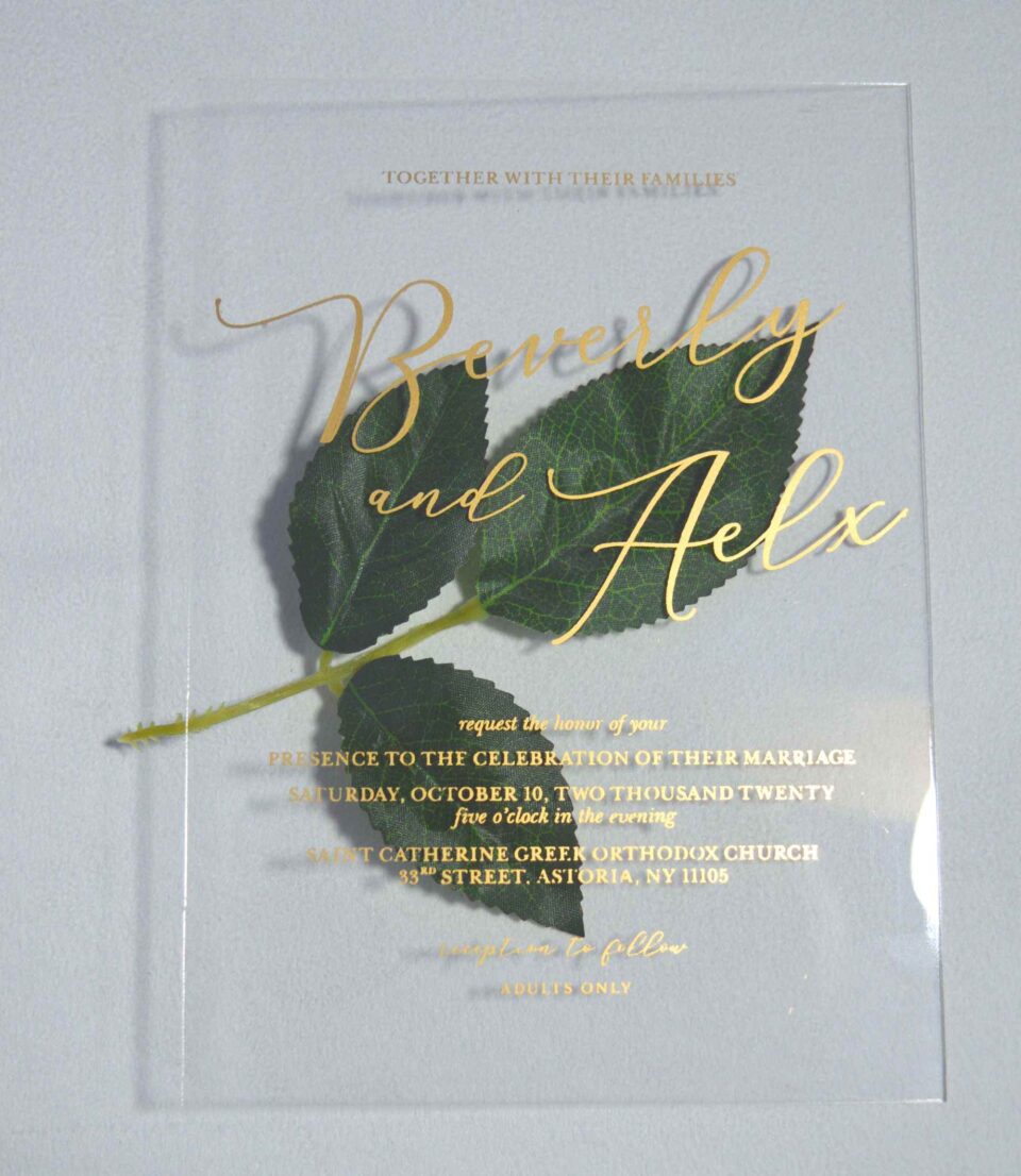 A transparent, rectangular acrylic wedding invitation is laid flat on a white surface, accompanied by a stem of green leaves at the center. The invitation features elegant gold text that casts subtle shadows onto the background.