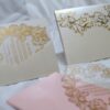 An ornate wedding invitation suite is displayed on a white surface, featuring ivory, pink, and transparent acrylic elements decorated with elaborate gold foil filigree.