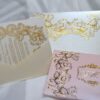 An ornate wedding invitation suite is displayed on a white surface, featuring ivory, pink, and transparent acrylic elements decorated with elaborate gold foil filigree.