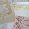 An ornate wedding invitation suite is displayed on a white surface, featuring ivory, pink, and transparent acrylic elements decorated with elaborate gold foil filigree.