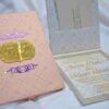 A high-angle shot featuring an open peach-colored wedding invitation box set on a white surface with draped white fabric and a gold lace cuff. On the left, the closed lid of the box displays a central gold plaque with the monogram "R & P," framed by purple scrollwork accented with pearls. On the right, the open box base reveals a formal acrylic invitation for "James Parker & Karen Brett" printed with gold text. A gold RSVP card with matching scrollwork is partially tucked into a pocket in the open lid.