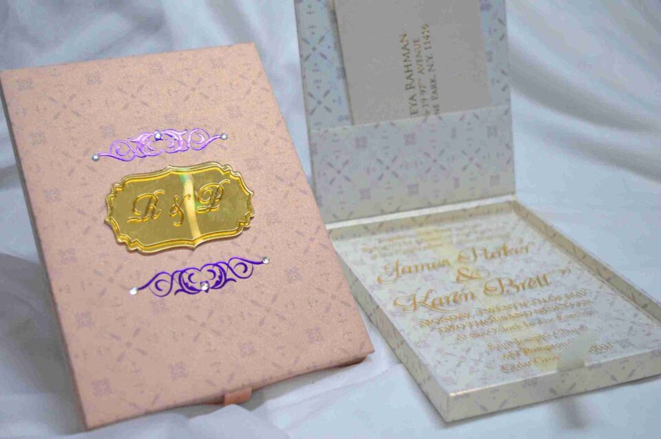 A high-angle shot featuring an open peach-colored wedding invitation box set on a white surface with draped white fabric and a gold lace cuff. On the left, the closed lid of the box displays a central gold plaque with the monogram "R & P," framed by purple scrollwork accented with pearls. On the right, the open box base reveals a formal acrylic invitation for "James Parker & Karen Brett" printed with gold text. A gold RSVP card with matching scrollwork is partially tucked into a pocket in the open lid.