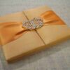 A rectangular, gold silk-textured gatefold invitation box shown from a perspective angle. A wide, shimmering orange satin ribbon wraps horizontally around the box, gathered at the center by an ornate, oval-shaped silver brooch encrusted with clear, sparkling crystals. The box rests on a neutral-toned, textured burlap or linen surface. The lighting emphasizes the fine, horizontal weave of the silk and the warm, metallic tones of the invitation suite.