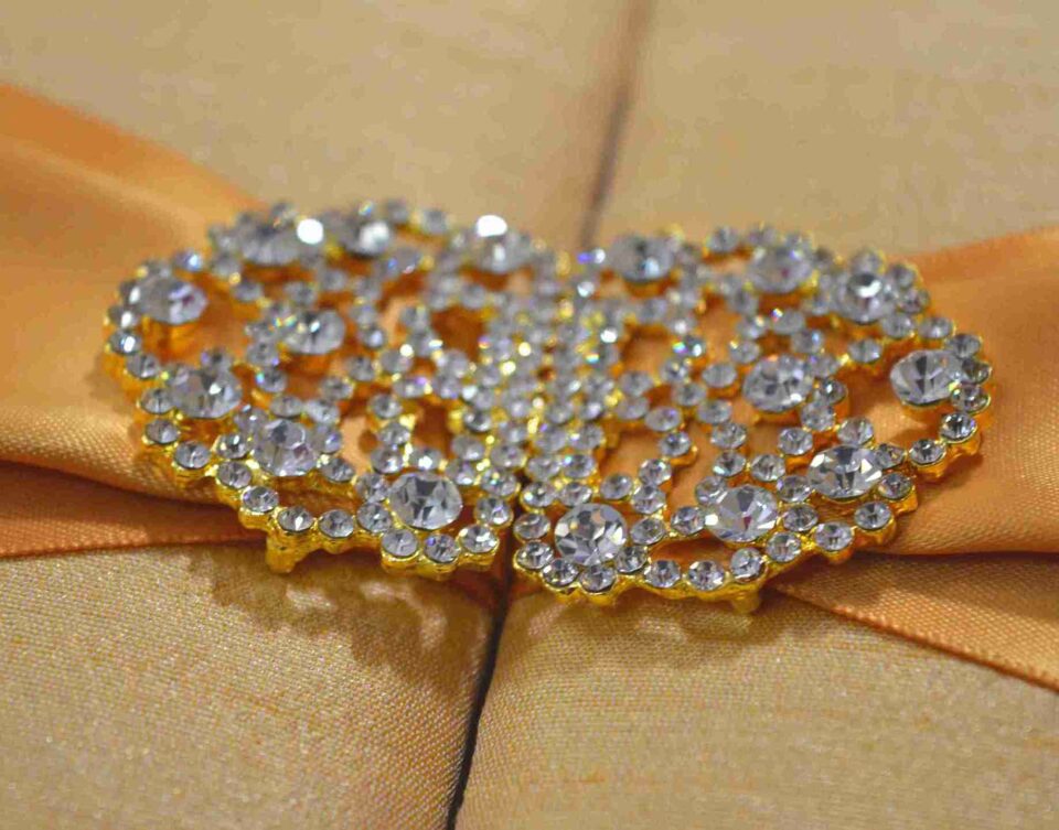 A rectangular, gold silk gatefold wedding invitation box shown from a perspective angle. A wide, shimmering gold satin ribbon wraps horizontally around the box, gathered at the center by an ornate, oval-shaped silver brooch encrusted with clear, sparkling crystals. The box rests on a neutral-toned, textured burlap or linen surface. The lighting emphasizes the fine, horizontal weave of the silk and the warm, metallic tones of the invitation suite.