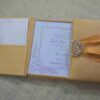 An open, gold silk-textured gatefold wedding invitation box resting on a burlap surface. The central panel displays the wedding invitation for "Lada Yana," featuring elegant cursive text and a delicate floral illustration in the corner. The left flap contains a pocket holding a white RSVP card with lace-like detailing. On the right, a shimmering orange satin ribbon is visible, secured by an ornate, crystal-encrusted silver brooch. The image is taken from a low side angle, highlighting the depth and texture of the luxury invitation suite.