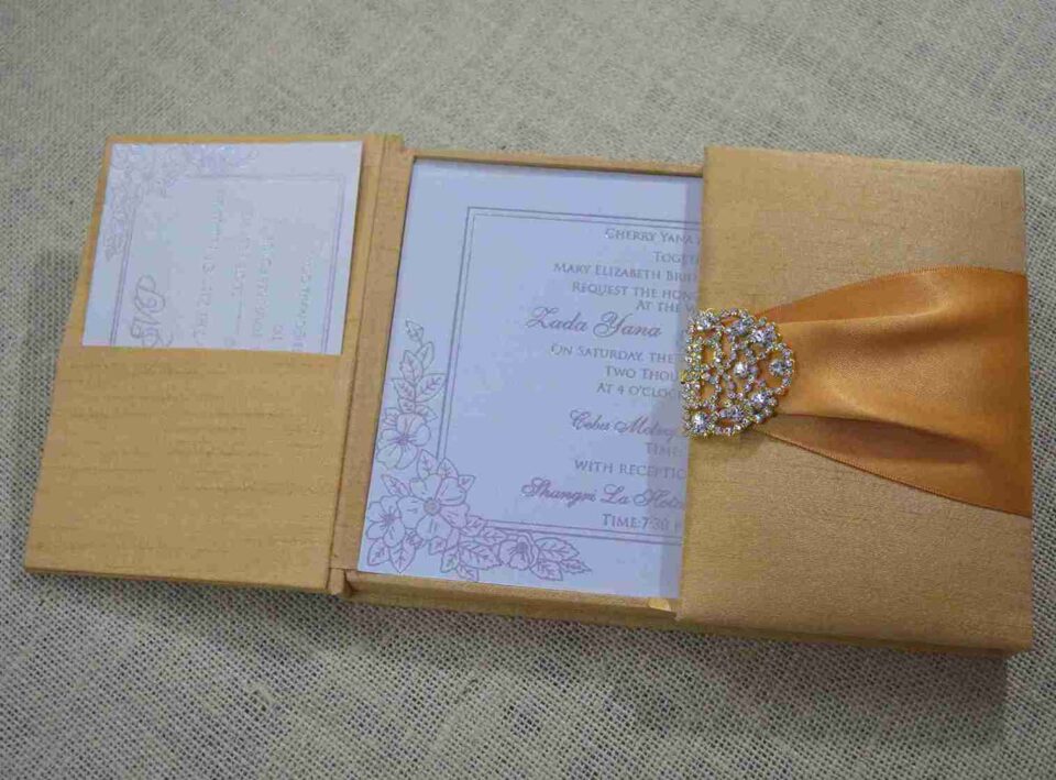An open, gold silk-textured gatefold wedding invitation box resting on a burlap surface. The central panel displays the wedding invitation for "Lada Yana," featuring elegant cursive text and a delicate floral illustration in the corner. The left flap contains a pocket holding a white RSVP card with lace-like detailing. On the right, a shimmering orange satin ribbon is visible, secured by an ornate, crystal-encrusted silver brooch. The image is taken from a low side angle, highlighting the depth and texture of the luxury invitation suite.