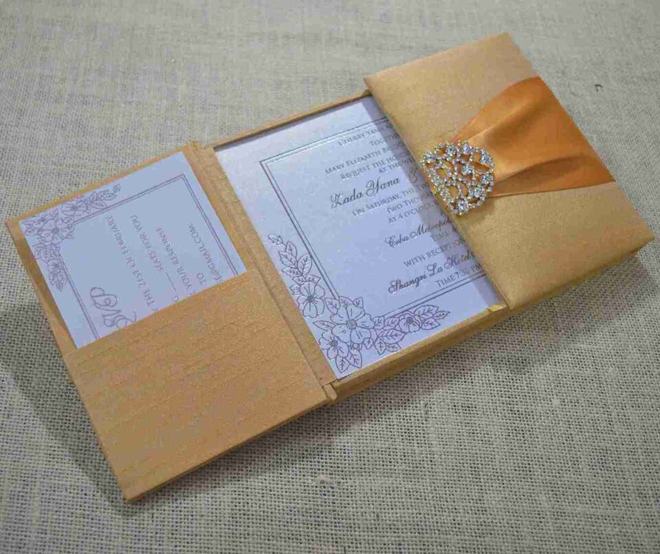 An open, gold silk-textured gatefold wedding invitation box resting on a burlap surface. The central panel displays the wedding invitation for "Lada Yana," featuring elegant cursive text and a delicate floral illustration in the corner. The left flap contains a pocket holding a white RSVP card with lace-like detailing. On the right, a shimmering orange satin ribbon is visible, secured by an ornate, crystal-encrusted silver brooch. The image is taken from a low side angle, highlighting the depth and texture of the luxury invitation suite.