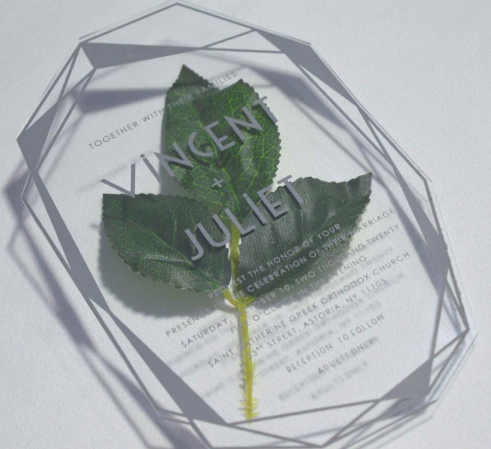 A clear acrylic wedding invitation in a geometric, diamond-like shape is positioned over a green leaf on a white surface. The invitation features the names in a large, bold sans-serif font, with the rest of the text in a smaller, lighter font. The text on the invitation read.
