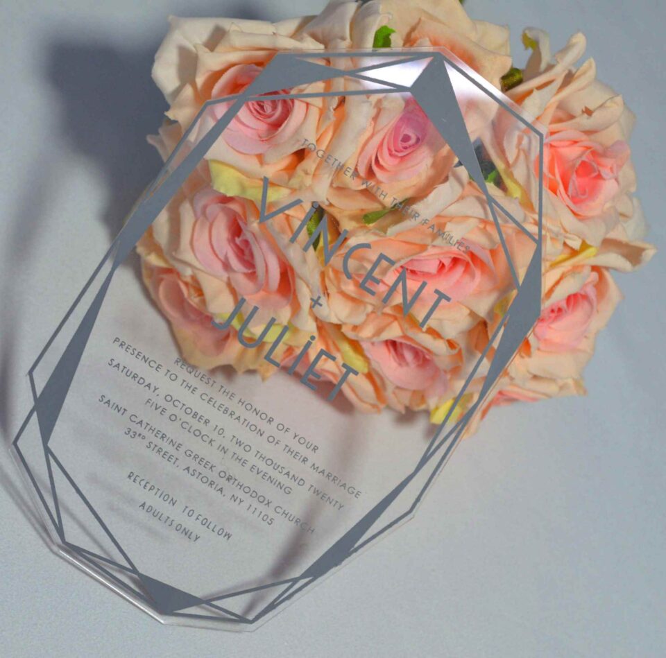 An angled shot of a transparent acrylic wedding invitation leaning against a bouquet of soft peach and pink roses. The invitation is uniquely shaped with a hexagonal, multi-faceted geometric border printed in a matte silver or grey ink.