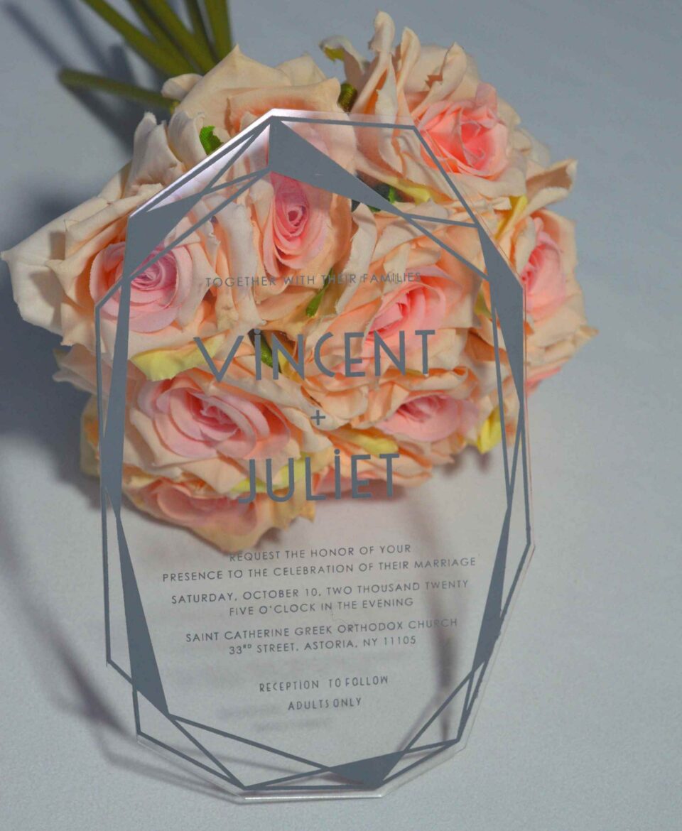 An angled shot of a transparent acrylic wedding invitation leaning against a bouquet of soft peach and pink roses. The invitation is uniquely shaped with a hexagonal, multi-faceted geometric border printed in a matte silver or grey ink.