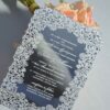This image features a high-end silver mirror acrylic wedding invitation with an intricate white laser-cut lace border. The card is shown at an angle, leaning against a bouquet of soft peach roses on a white surface. The invitation features elegant white printed text with the details.