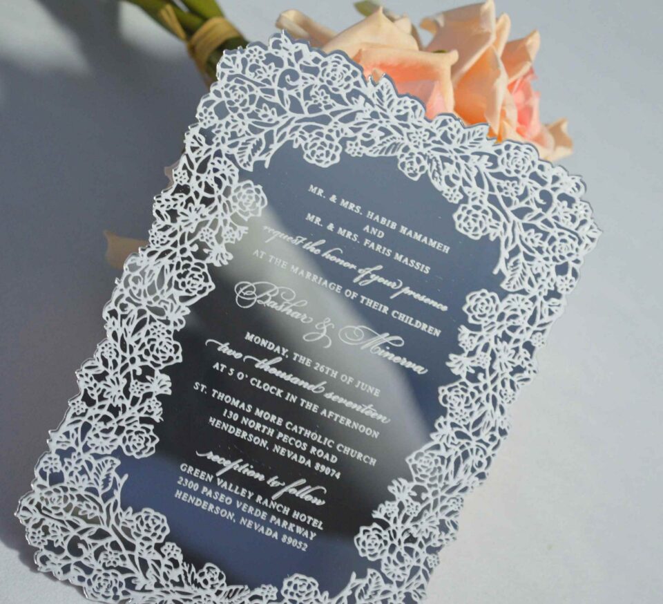 This image features a high-end silver mirror acrylic wedding invitation with an intricate white laser-cut lace border. The card is shown at an angle, leaning against a bouquet of soft peach roses on a white surface. The invitation features elegant white printed text with the details.