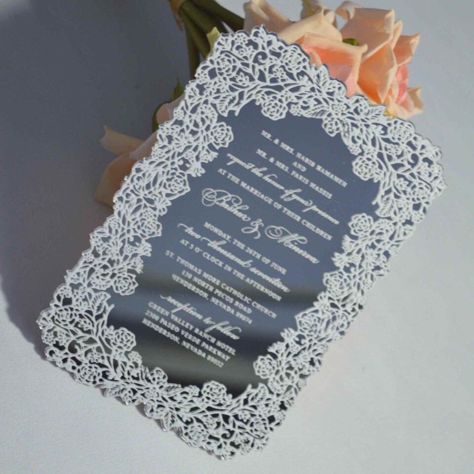 This image features a high-end silver mirror acrylic wedding invitation with an intricate white laser-cut lace border. The card is shown at an angle, leaning against a bouquet of soft peach roses on a white surface. The invitation features elegant white printed text with the details.